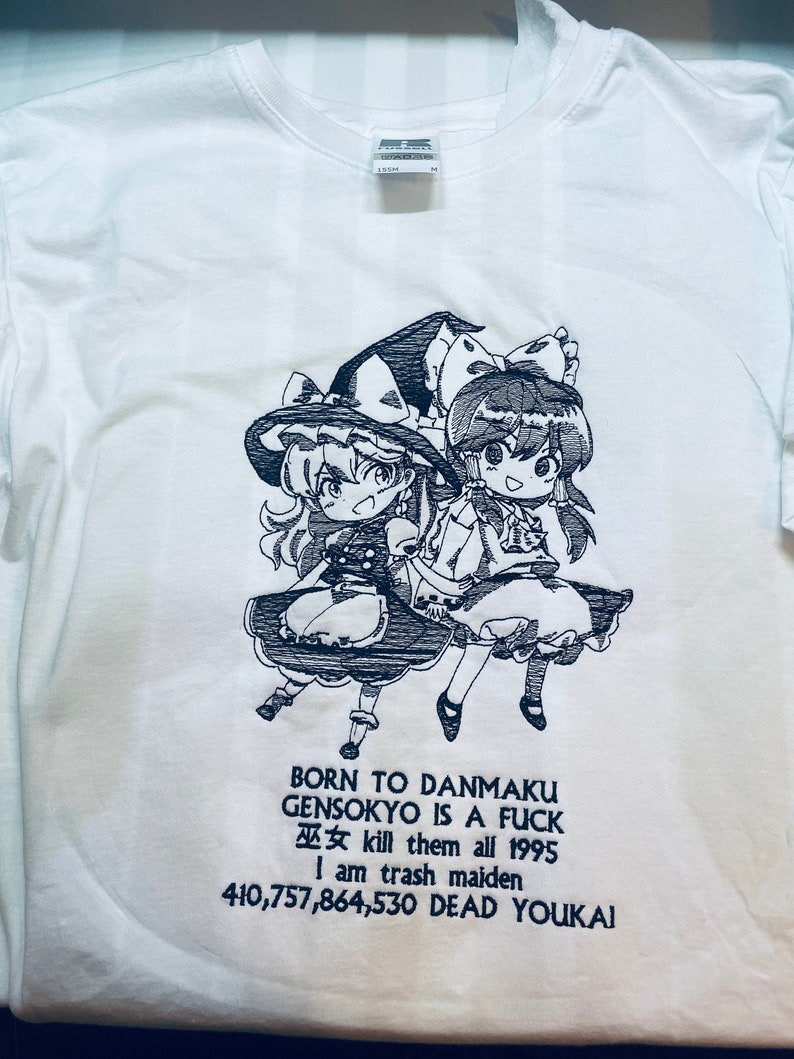 Pu&ograve; includere: Maglietta bianca con un'illustrazione in bianco e nero di due personaggi anime. Il testo sulla maglietta recita "BORN TO DANMAKU GENSOKYO IS A FUCK 巫女 kill them all 1995 I am trash maiden 410,757,864,530 DEAD YOUKAI".