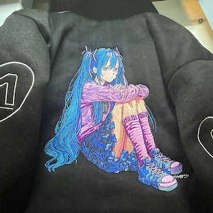 Puede incluir: Sudadera negra con una imagen bordada de una chica de anime de pelo azul. Lleva una camisa morada y una falda negra, y est&aacute; sentada con las piernas cruzadas y los brazos cruzados. Lleva calcetines rosas y zapatillas negras y blancas.
