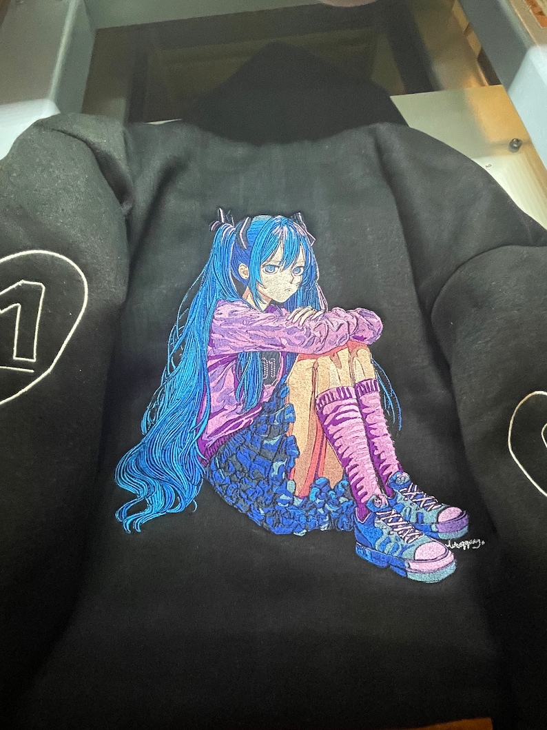 Puede incluir: Sudadera negra con una imagen bordada de una chica de anime de pelo azul. Lleva una camisa morada y una falda negra, y est&aacute; sentada con las piernas cruzadas y los brazos cruzados. Lleva calcetines rosas y zapatillas negras y blancas.