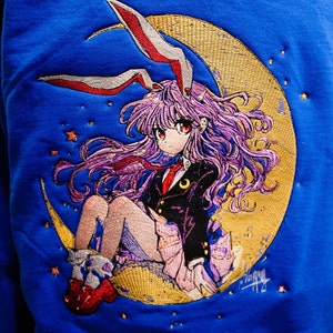 May include: Embroidered design on a blue sweatshirt featuring a girl with purple hair wearing a black and red outfit, sitting on a crescent moon with stars.