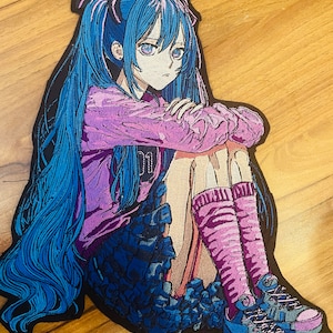 Pode incluir: Um grande emblema bordado apresentando uma personagem estilo anime com longos cabelos azuis em maria-chiquinhas, vestindo uma camisa roxa de manga comprida com o número 01, uma saia azul escura, meias roxas até o joelho e tênis azuis e roxos.
