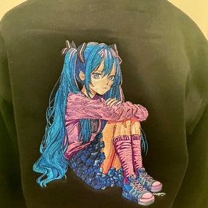 Puede incluir: Sudadera negra con una imagen bordada de un personaje de anime con cabello azul, vistiendo una camisa rosa y una falda azul, sentada con las piernas cruzadas. El personaje tiene una expresi&oacute;n triste en su rostro.
