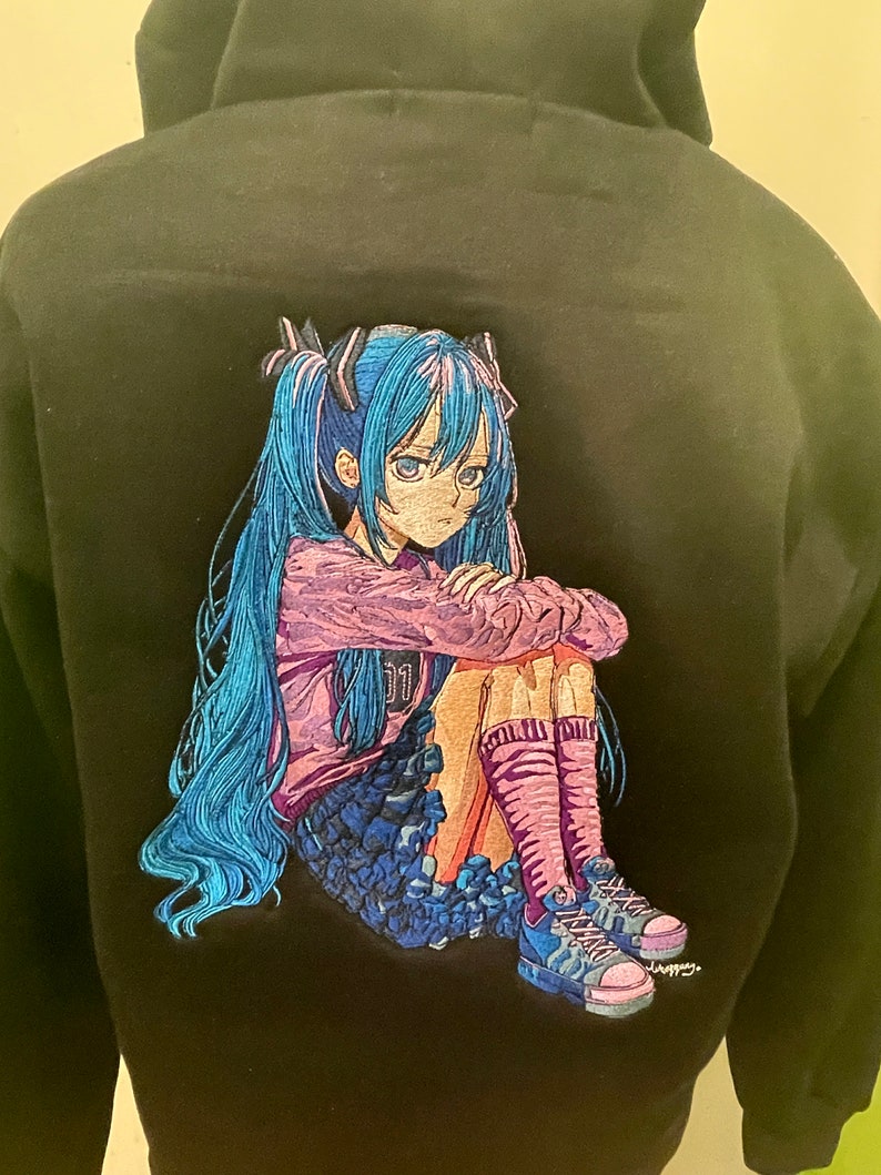 Puede incluir: Sudadera negra con una imagen bordada de un personaje de anime con cabello azul, vistiendo una camisa rosa y una falda azul, sentada con las piernas cruzadas. El personaje tiene una expresi&oacute;n triste en su rostro.