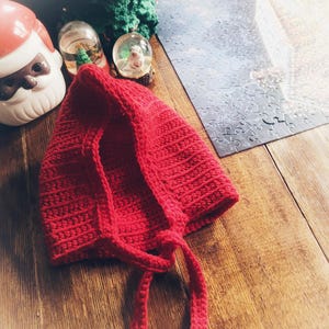 May include: A handmade, bright red crochet bonnet with long ties. The bonnet is made of a textured yarn and is likely for a baby or child. The background includes Christmas decorations, such as a Santa Claus figurine, snow globes, and a green crochet Christmas tree.
