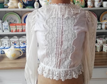 May include: A white lace blouse with long sleeves and a high collar. The blouse features a delicate lace trim around the collar, cuffs, and hem. The lace is a floral pattern and the blouse is made of a sheer fabric.