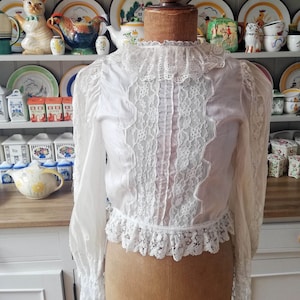 May include: A white lace blouse with long sleeves and a high collar. The blouse has a delicate lace trim around the collar, cuffs, and hem. The lace is a floral pattern and the blouse is made of a sheer fabric.
