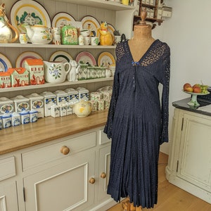 May include: A navy blue lace dress with a V-neckline and long sleeves. The dress has a pleated skirt and a button closure at the neckline. The dress is displayed on a mannequin in a kitchen setting.