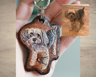 May include: A brown leather keychain with a painted portrait of a Yorkshire Terrier. The keychain has a silver metal ring and clasp. The dog is brown and black with a white chest and paws.