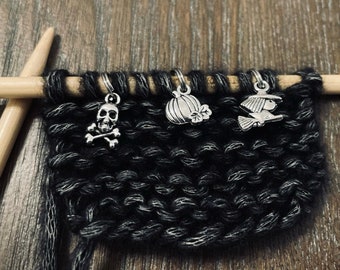 May include: Three silver Halloween-themed stitch markers on black knitting needles. The markers are a skull and crossbones, a pumpkin, and a witch on a broomstick.