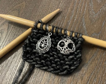 May include: Close-up of a black knitted fabric with two silver charms attached. The charms are a spider web with a spider and a skull with floral details. The fabric is on a pair of wooden knitting needles.