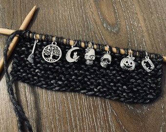 May include: A set of eight Halloween-themed knitting stitch markers, including a broom, a tree, a crescent moon, a cat, a skull with glasses, a skull, a jack-o'-lantern, and a set of fangs. The markers are attached to a black knitting project.