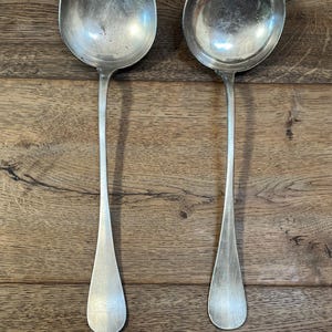May include: A pair of antique silver ladles, each with a long handle and a round bowl. The ladles are placed on a wooden surface, highlighting their polished, reflective finish. These spoons are likely for serving soups or sauces.