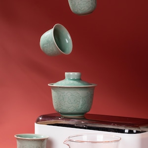 Puede incluir: Un juego de té de cerámica celadón con diseños florales, que incluye tazas y un gaiwan con tapa. También hay una jarra de vidrio con té y una caja blanca y marrón, todo sobre un fondo rojo.