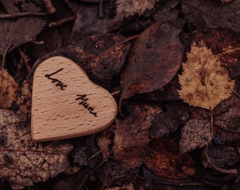 May include: A wooden heart with the words "Love Mum" engraved in black script, resting on a bed of fallen leaves. The heart is light brown, contrasting with the darker shades of the leaves.