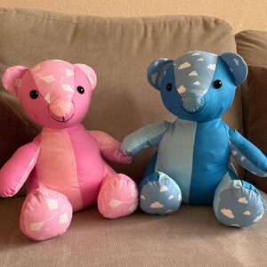 May include: Two handmade plush teddy bears sitting on a grey sofa. The bear on the left is pink with a white kite pattern and the bear on the right is blue with a white cloud pattern.