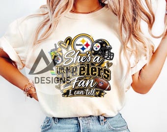 May include: A cream-colored t-shirt featuring a graphic print with a Pittsburgh Steelers helmet, a football, a tumbler, and hearts. The text reads "She's a Steelers Fan I can tell" in black and gold lettering, with a small NFL logo on the football.
