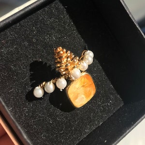 Peut inclure: Broche dorée avec un motif de pomme de pin, de petites perles blanches et une pierre carrée orange. La broche est présentée dans un écrin noir. La lumière se reflète sur l'or et les perles.