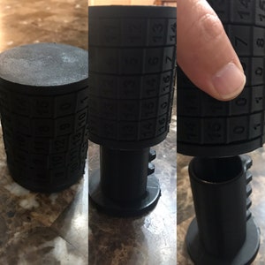 Puede incluir: Una cerradura de combinación negra con diales numerados. La cerradura está hecha de plástico y tiene una forma cilíndrica. La cerradura está en posición abierta, mostrando los números en los diales.