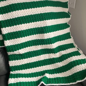 May include: A green and white striped crocheted blanket with a textured pattern. The blanket is folded and draped over a couch.