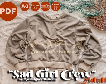 Może przedstawiać: Beżowa bluza z okrągłym dekoltem z nadrukiem "Sad Girl Crew" i "Nostalgia's Patterns" z przodu. Bluza leży na białej i brązowej, teksturowanej powierzchni.