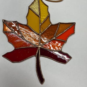 May include: A stained glass maple leaf ornament with a copper border. The leaf features a gradient of colors, including yellow, orange, and red. A brown cord is attached for hanging. This autumn-themed decoration is ideal for fall.