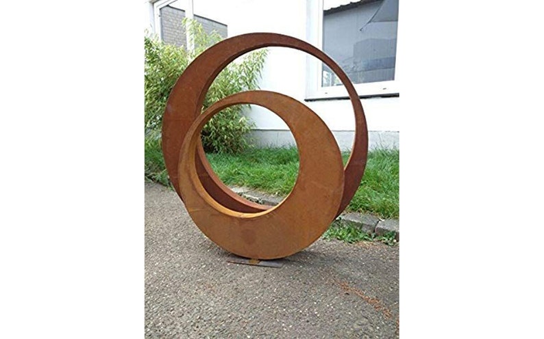 May include: A rusty metal sculpture of two overlapping circles, one larger than the other, standing on a concrete surface. The sculpture is outdoors in a garden setting.