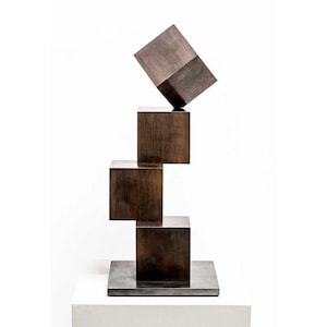 Op de afbeelding: Een sculptuur gemaakt van vier gestapelde bronzen kubussen. De bovenste kubus is schuin geplaatst. De sculptuur staat op een vierkante basis.