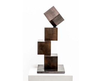 Könnte beinhalten: Eine Skulptur aus vier übereinander gestapelten Bronzewürfeln. Der oberste Würfel ist schräg gestellt. Die Skulptur steht auf einem quadratischen Sockel.