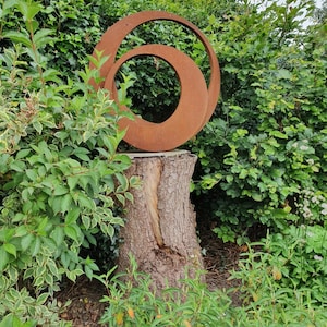 May include: A rusty metal sculpture of two overlapping circles, mounted on a tree stump in a garden setting.