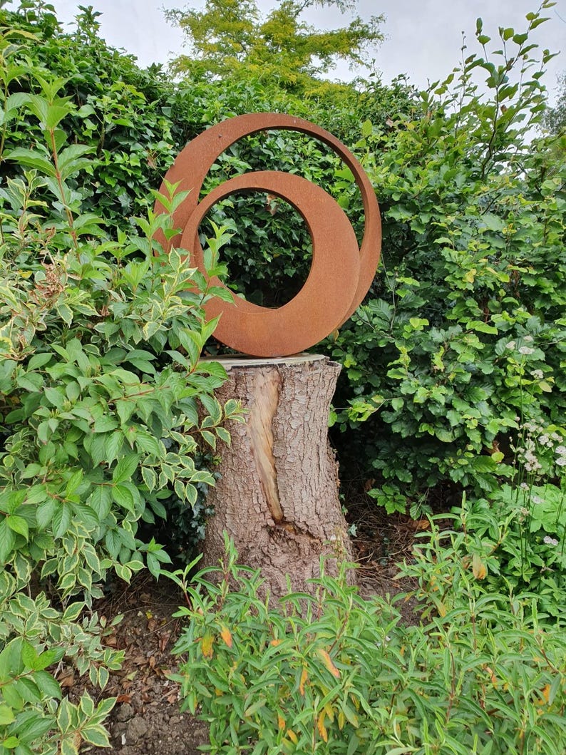 May include: A rusty metal sculpture of two overlapping circles, mounted on a tree stump in a garden setting.