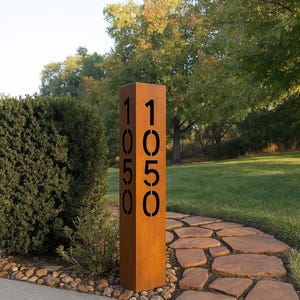 Può includere: Un'insegna indirizzo rettangolare in metallo usurato con il numero "1050" intagliato in nero. L'insegna marrone è in un giardino, vicino a un sentiero di pietra e fogliame verde.
