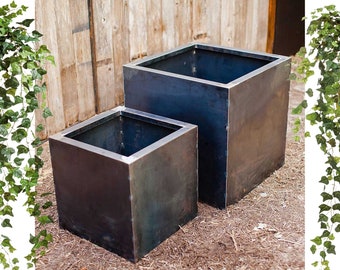 May include: Two square metal planters, one larger than the other, are placed on a bed of brown mulch. The planters are a dark grey colour with a slightly rusted finish.