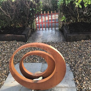 May include: A rusty metal sculpture of two overlapping circles in a garden setting. The sculpture is on a gray stone patio surrounded by gravel.