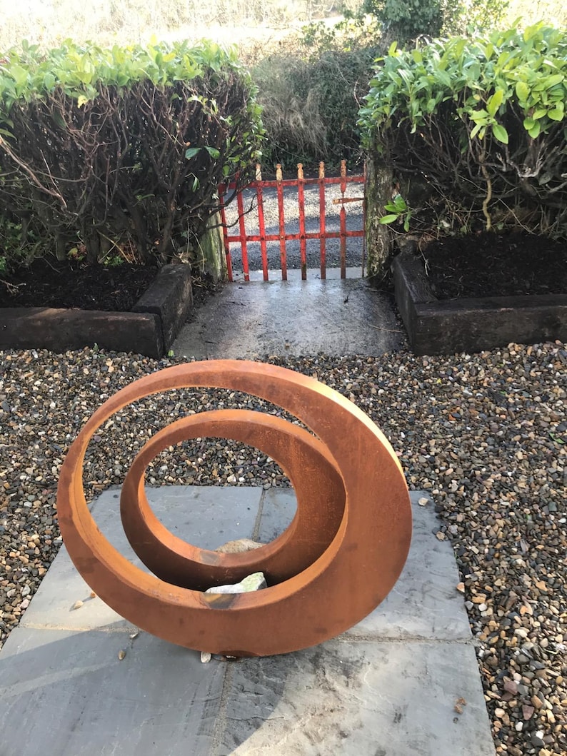 May include: A rusty metal sculpture of two overlapping circles in a garden setting. The sculpture is on a gray stone patio surrounded by gravel.