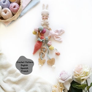 May include: A crocheted baby toy with a wooden ring, a carrot, a bunny, and a heart. The toy is made in pastel colors and has a variety of textures. The toy is on a white background with a blanket and flowers.