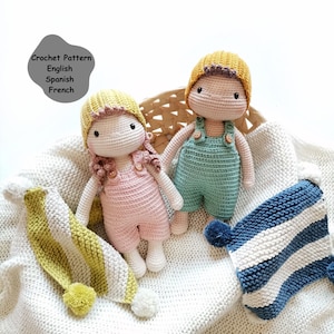 Könnte beinhalten: Zwei gehäkelte Puppen, die Overalls und Hüte tragen. Die Mädchenpuppe trägt ein rosa Outfit und einen gelben Hut. Die Jungenpuppe trägt ein grünes Outfit und einen gelben Hut. Die Puppen sitzen auf einer weißen Decke mit einer blau-weiß gestreiften Decke und einer gelb-weiß gestreiften Decke.