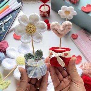 Puede incluir: Decoraciones de flores y corazones de ganchillo en macetas pequeñas. La margarita blanca y el corazón rosa están sobre palitos de madera. Las macetas son rojas y azules, con detalles de cintas y lazos. Pequeños corazones de ganchillo dispersos.