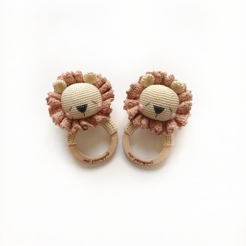 May include: Two crochet lion rattles featuring beige heads with dark embroidered eyes and noses, pink cheeks, and ruffled dusty pink manes. Each lion is attached to a smooth, light-colored wooden ring engraved with the text mio pineapple.
