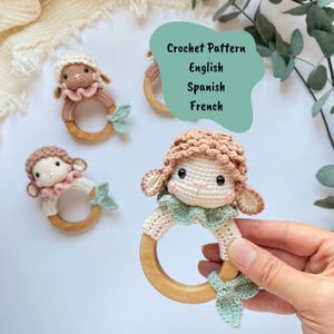 May include: Crocheted sheep baby toys with wooden rings. The sheep have cream-coloured bodies, pink and brown accents, and light green leaf-shaped details. The text "Crochet Pattern English Spanish French" is visible.