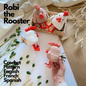 Op de afbeelding: Vier handgemaakte gehaakte haan-speeltjes in beige en wit met rode kammen en lellen. Twee hanen zitten in een klein gevlochten mandje, terwijl er twee buiten liggen. De tekst luidt Robi the Rooster Crochet Pattern English French Spanish.