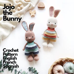 May include: Two crocheted bunny dolls labeled Jojo the Bunny. One bunny is brown with blue-striped shorts, the other is cream with a multi-colored striped dress. The text reads Jojo the Bunny Crochet Pattern English French Spanish.