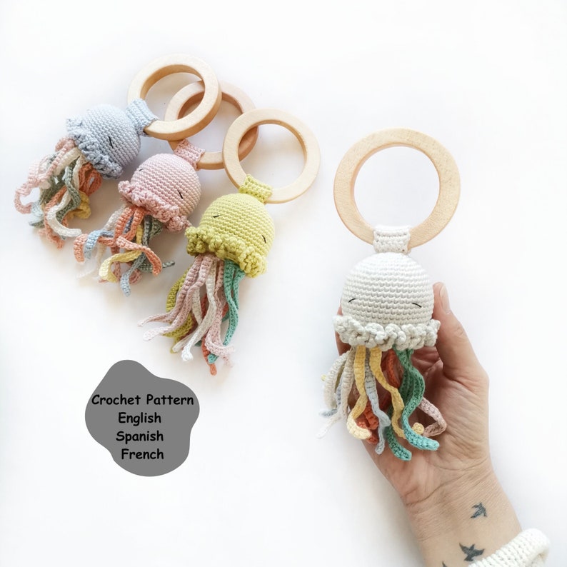 May include: Four crochet jellyfish toys attached to smooth wooden rings. Each jellyfish features a rounded head with embroidered closed eyes and dangling multi-colored tentacles in shades of blue, pink, yellow, and white. Text reads: Crochet Pattern English Spanish French.