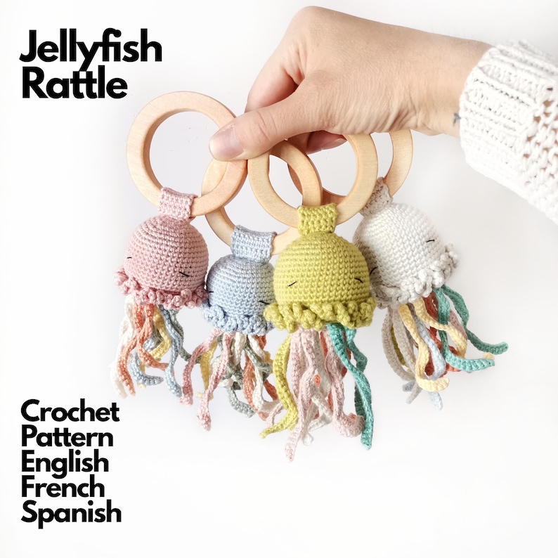 May include: Four jellyfish rattles featuring crocheted heads in dusty pink, light blue, olive green, and cream, each attached to a smooth wooden ring. The jellyfish have embroidered closed eyes and dangling multi-colored yarn tentacles.