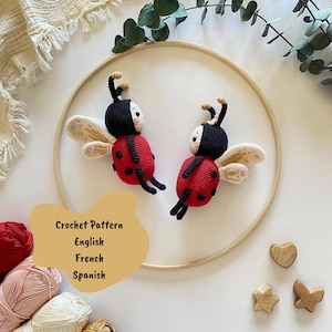 Könnte beinhalten: Zwei handgefertigte Häkel-Marienkäfer mit roten Körpern, schwarzen Punkten und schwarz-weißen Gesichtern. Sie haben beige Flügel und sind auf einem Holzreifen platziert. Das Bild enthält den Text "Crochet Pattern" auf Englisch, Französisch und Spanisch.