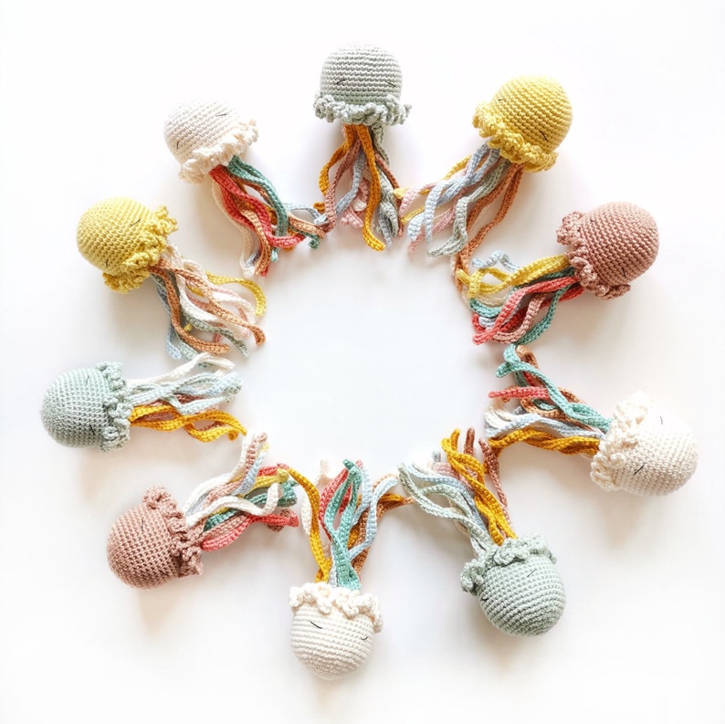May include: A circular arrangement of ten small, crocheted jellyfish toys. Each jellyfish features a rounded, solid-colored head in shades of cream, yellow, sage green, or dusty pink, with multiple dangling, multi-colored yarn tentacles.