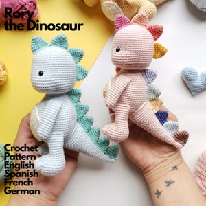 Könnte beinhalten: Zwei gehäkelte Dinosaurier-Spielzeuge, eines hellblau und eines hellrosa, jeweils mit einem cremefarbenen Bauch und einer Reihe bunter Stacheln auf dem Rücken. Der Text Rory the Dinosaur Crochet Pattern English Spanish French German ist sichtbar.