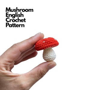 Puede incluir: Un pequeño hongo de ganchillo con un sombrero rojo con puntos blancos y un tallo de color crema. El texto Mushroom English Crochet Pattern es visible en la esquina superior izquierda. El objeto se sostiene entre los dedos sobre un fondo blanco.