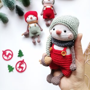 May include: A handcrafted crochet snowman doll wearing a sage green knit hat, a dark gray scarf, and red overalls with two small wooden buttons. The doll has a white round head, a small orange nose, black bead eyes, and pink blush accents.