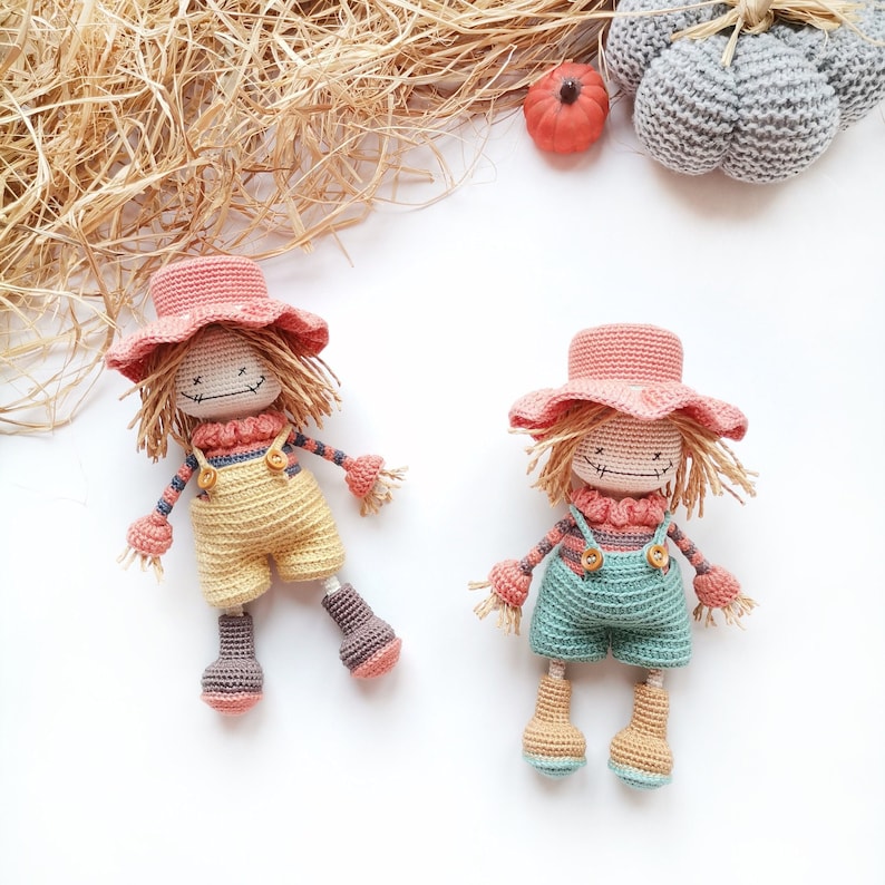 May include: Two handmade crochet scarecrow dolls featuring peach-colored hats, straw-like hair, and stitched facial features. One doll wears yellow overalls, while the other wears green overalls. Both dolls have striped long-sleeve shirts and boots.