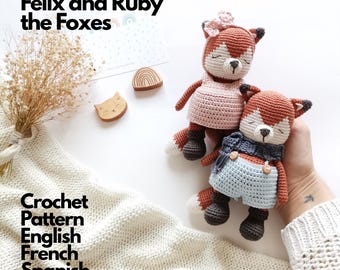 Op de afbeelding: Twee handgemaakte gehaakte vossenpoppen genaamd Felix en Ruby. De ene vos draagt een lichtroze jurk met een bijpassende bloem, terwijl de andere een lichtblauwe tuinbroek en een donkerblauwe sjaal draagt. Beide hebben witte gezichten en donkerbruine laarzen.